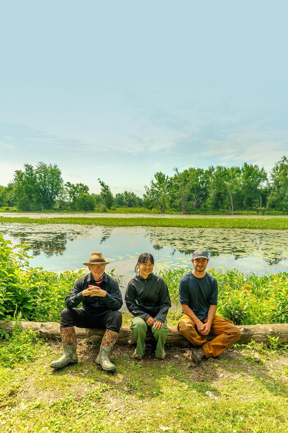 Trois personnes, deux hommes et une femme, sont assises sur un tronc d’arbre devant une étendue d’eau.